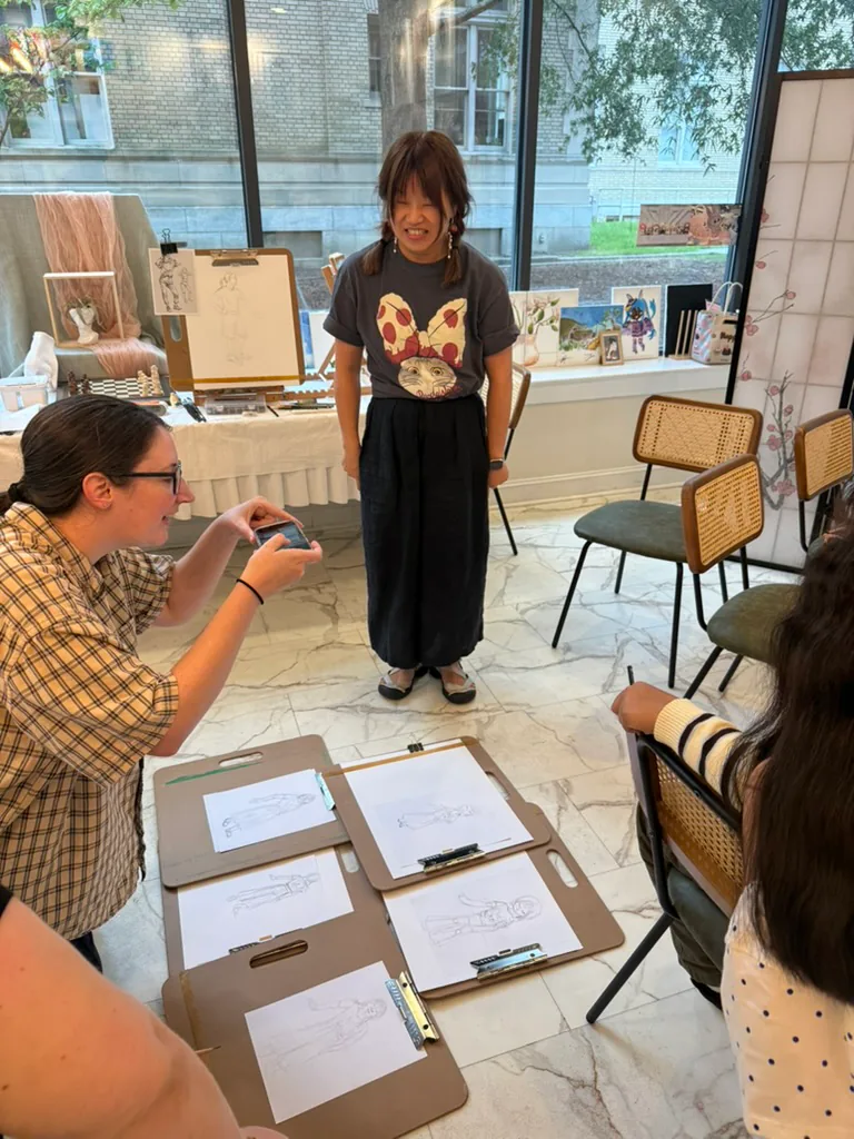 A clothed participant poses in the center while seated sketchers work from clipboards.