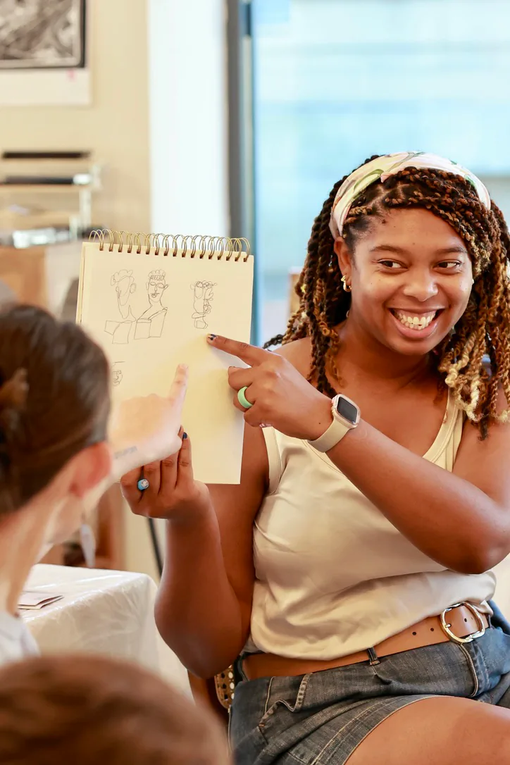A smiling Sketch Club participant holds up a spiral sketchbook while another participant points to one of the drawings.