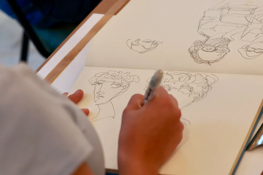 A hand draws a portrait in an open sketchbook filled with face studies.